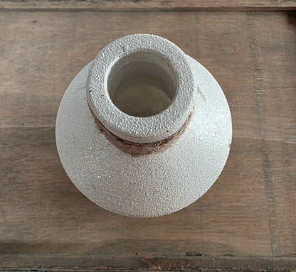 A handmade glass bottle with a textured, speckled off-white glaze and a rustic brown clay band around the neck, shown on a wooden surface.