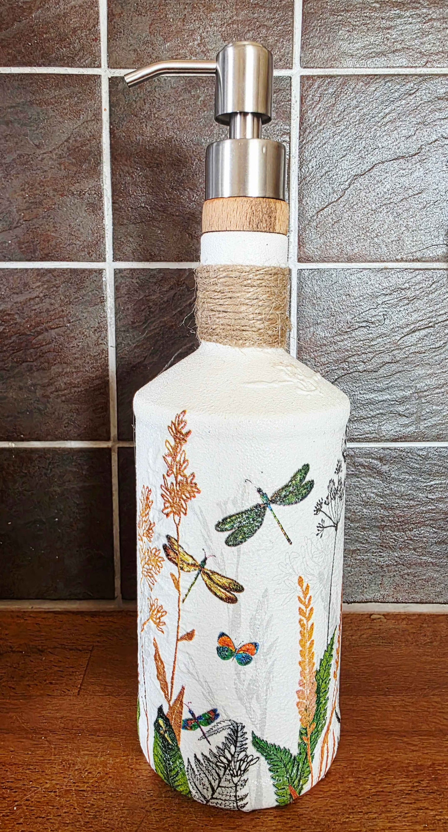 A decorative white glass soap dispenser bottle with a silver metal pump and a light wood collar, wrapped in twine near the neck. The bottle is decorated with hand-painted dragonflies, butterflies, and wildflowers.