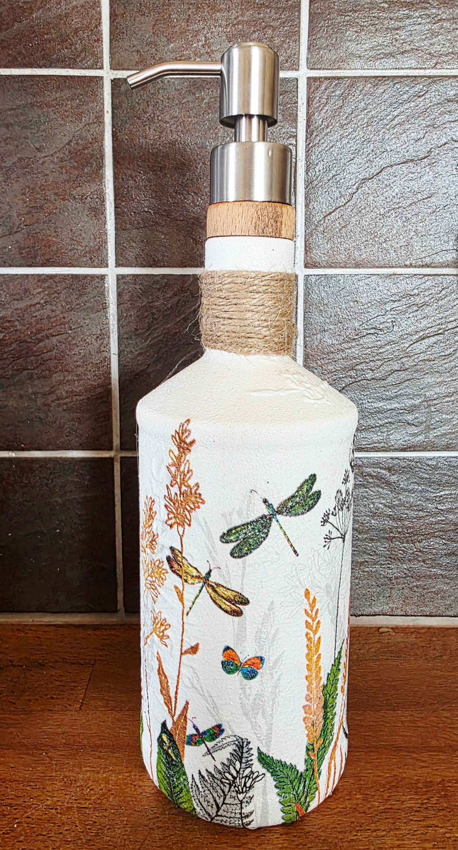 A decorative white glass soap dispenser bottle with a silver metal pump and a light wood collar, wrapped in twine near the neck. The bottle is decorated with hand-painted dragonflies, butterflies, and wildflowers.